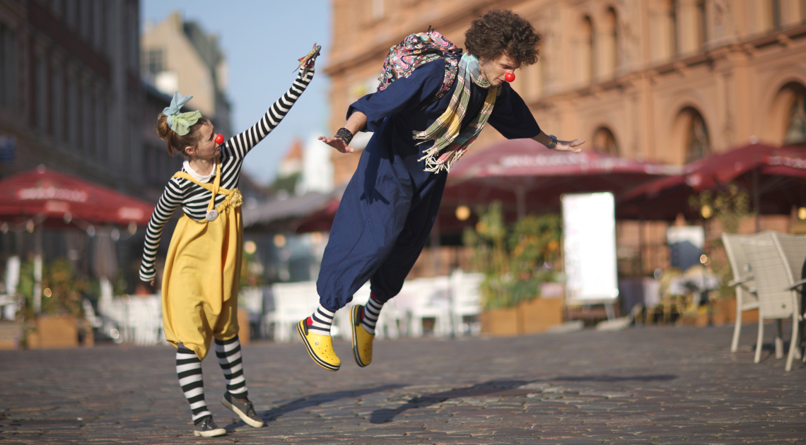 Kunst und Kultur im Aufwind: Ein Clownspaar stellt in einer Fußgängerzone pantomimisch ein Flugzeug nach